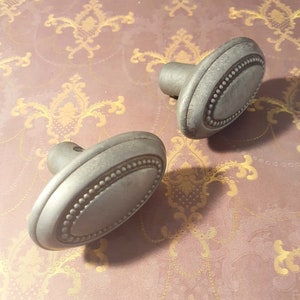 May include: Two antique-style, oval door knobs with a silver finish. Each knob has a beaded border around the central oval. The knobs are made of metal and are designed for doors or drawers.