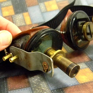 May include: Steampunk goggles with brass accents and a leather strap. The goggles have a round lens on one side and a telescope lens on the other.