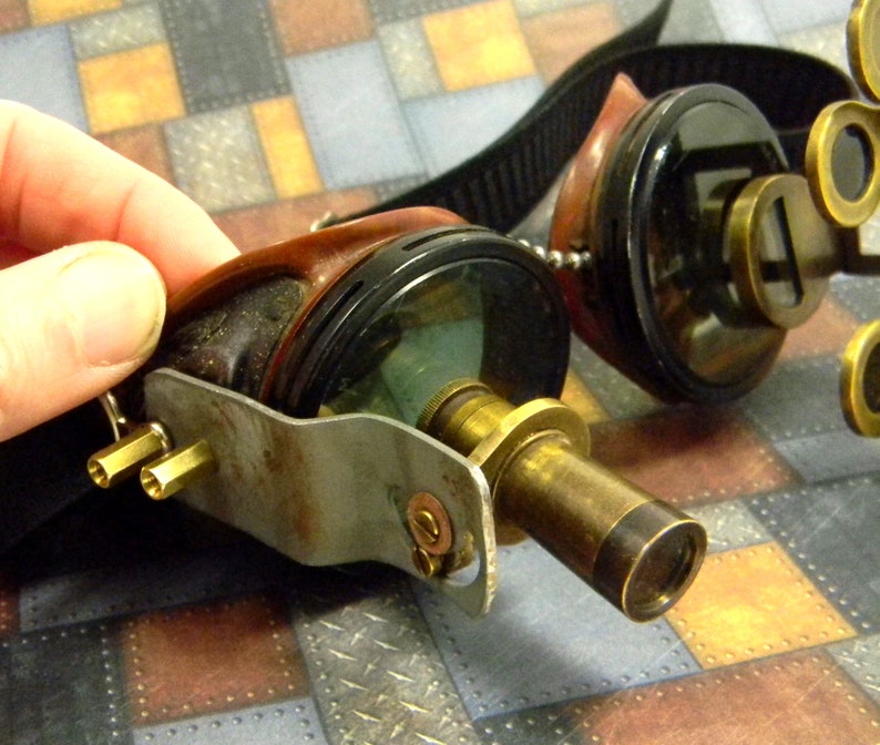 May include: Steampunk goggles with brass accents and a leather strap. The goggles have a round lens on one side and a telescope lens on the other.