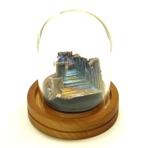 May include: A large, iridescent bismuth crystal is displayed in a glass dome on a wooden base. The crystal has a layered, geometric structure with a rainbow of colors.