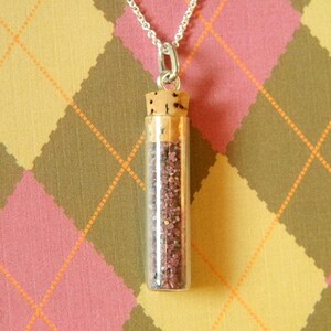 Può includere: Una collana con catena d'argento con un piccolo ciondolo di vetro a forma di fiala riempito di sabbia rosa e rossa. La fiala ha un tappo di sughero ed è sospesa dalla catena.