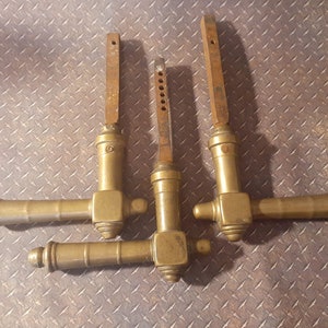 May include: Four antique brass door handles with a weathered appearance. Each handle has a horizontal grip and a vertical, rectangular extension. The handles are arranged on a textured, diamond-plate metal surface.