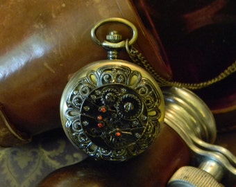 May include: A gold pocket watch with a black face and intricate details. The watch has a chain attached to it and is sitting on a brown leather surface.