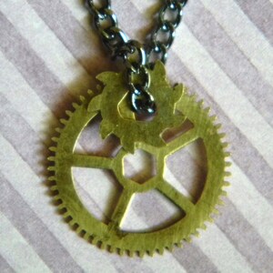 May include: A gold-toned gear necklace with a black chain. The gear has a detailed design with multiple spokes and a smaller gear in the center.