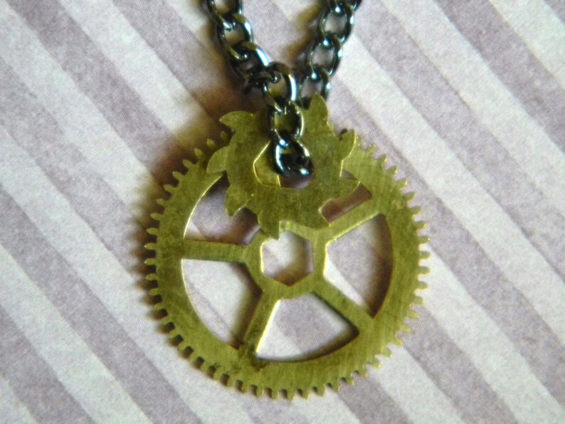 May include: A gold-toned gear necklace with a black chain. The gear has a detailed design with multiple spokes and a smaller gear in the center.
