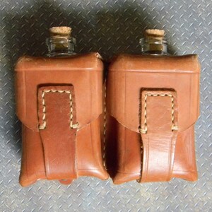 May include: Two brown leather pouches with stitched detailing, each containing a clear glass bottle with a cork stopper.