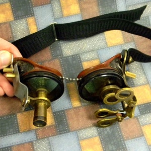 May include: A pair of steampunk goggles with a black adjustable strap, brass lenses, and a brown leather frame. The goggles have a chain connecting the two lenses.