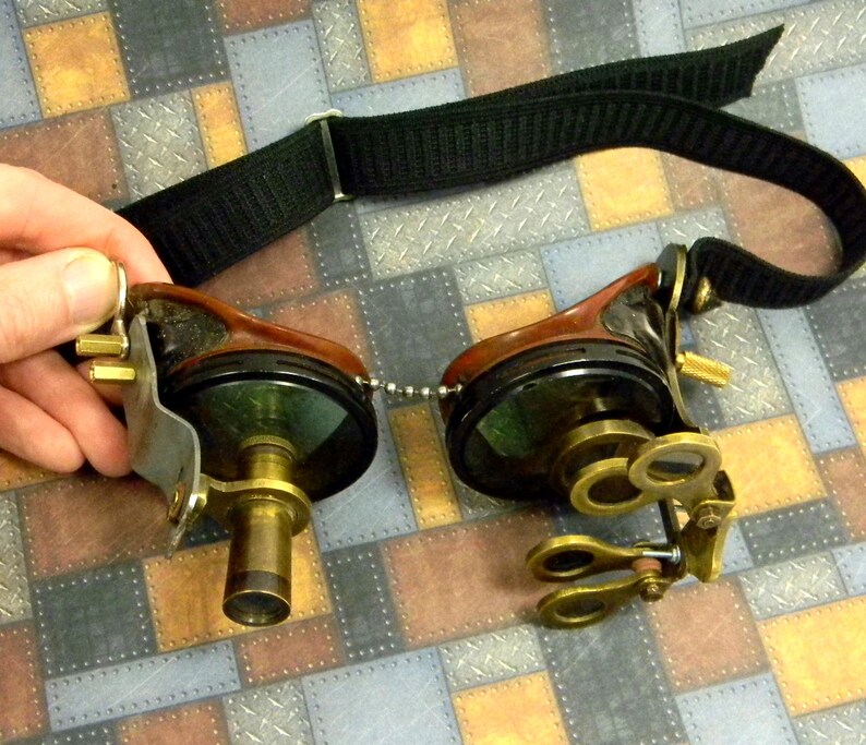 May include: A pair of steampunk goggles with a black adjustable strap, brass lenses, and a brown leather frame. The goggles have a chain connecting the two lenses.