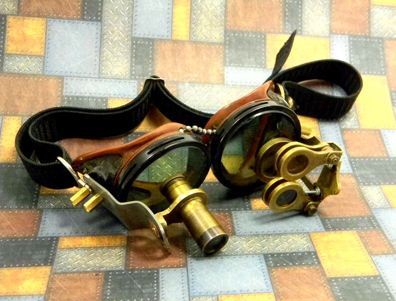 May include: A pair of steampunk goggles with a black leather strap, brown leather accents, and brass lenses. The goggles have a magnifying lens on the side.
