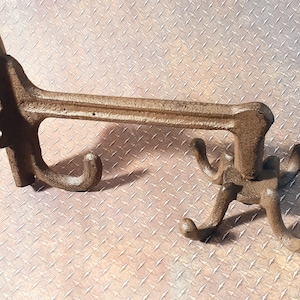 May include: A rustic cast iron wall hook with a single hook on one side and four hooks on the other side. The hook is a dark brown color and has a distressed finish.