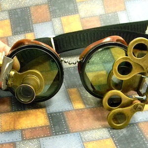 May include: Steampunk goggles with brass accents and adjustable lenses. The goggles have a black leather strap and a chain connecting the lenses.