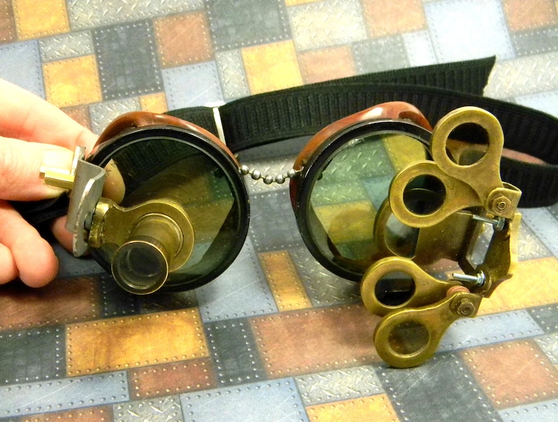 May include: Steampunk goggles with brass accents and adjustable lenses. The goggles have a black leather strap and a chain connecting the lenses.