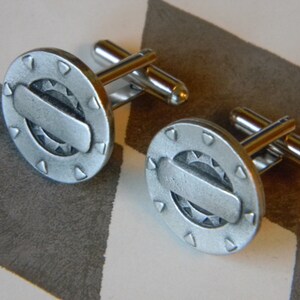 May include: A pair of silver cufflinks with a circular design featuring a gear and a rectangular shape in the centre. The cufflinks have a polished finish.