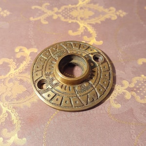 Pode incluir: Uma roseta de maçaneta de porta de latão com um design geométrico decorativo. A roseta tem forma circular com um furo central para a maçaneta.