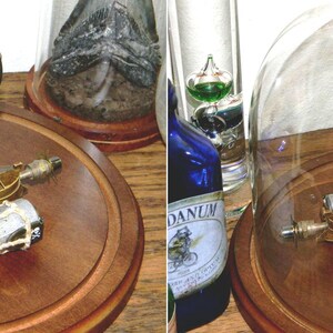 May include: A steampunk-style miniature airship model with a black balloon, brass-colored gondola, and silver cylinders, displayed on a wooden base. The model is under a glass dome, showcasing intricate details and a vintage aesthetic.