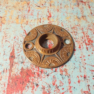 Pode incluir: Uma maçaneta de porta decorativa de latão com um design circular que apresenta padrões florais intrincados. A maçaneta tem um furo central para uma pega de porta.