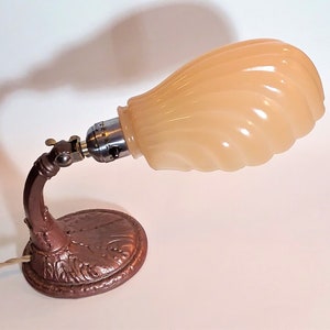 May include: A vintage desk lamp with a peach-colored, swirled glass shade. The lamp has a bronze-colored, ornate base and a curved arm. The lamp is plugged in and ready to use. The lamp is a decorative lighting fixture.