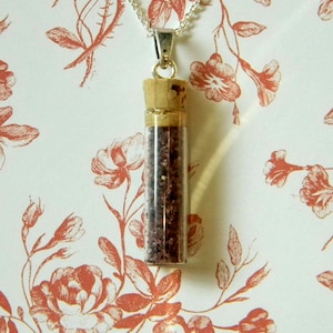 May include: A silver chain necklace with a cork-topped glass vial filled with small, dark red and brown particles. The vial is suspended from the chain and hangs against a white background with a floral pattern.