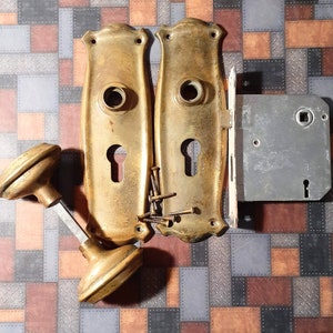 May include: Two antique brass door knobs with keyholes and a matching brass door latch. The knobs have a rounded top and a decorative design. The latch is rectangular with a square keyhole.