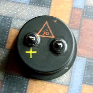 Puede incluir: Un componente el&eacute;ctrico redondo negro con dos terminales y un signo m&aacute;s amarillo. Un tri&aacute;ngulo con borde naranja y el n&uacute;mero 70 en su interior es visible en el componente.