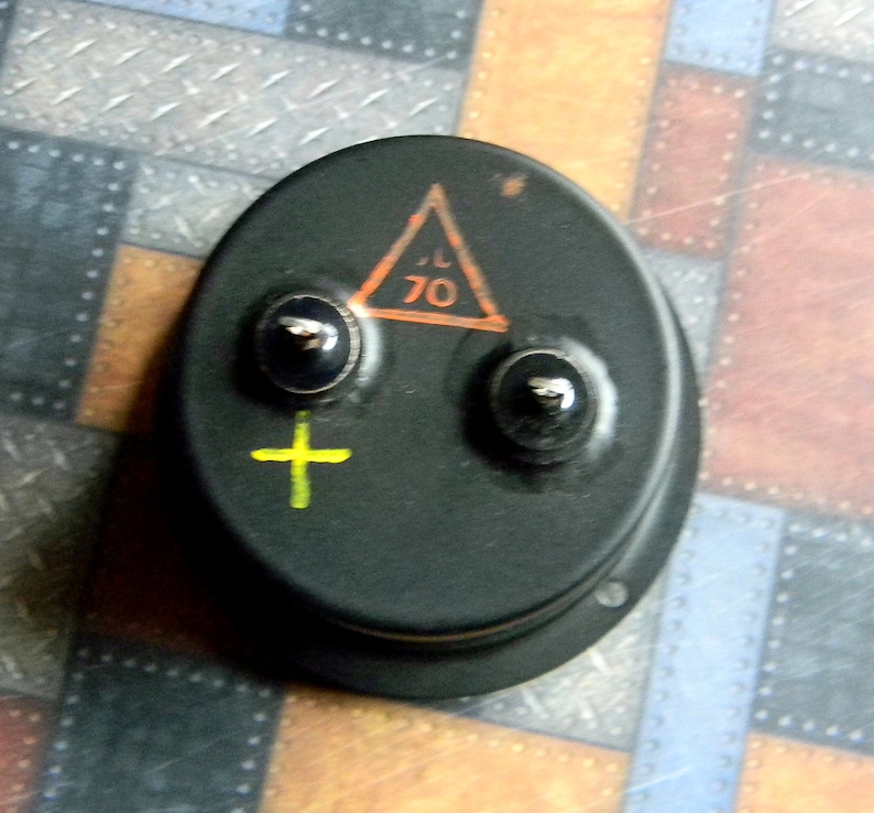 Puede incluir: Un componente el&eacute;ctrico redondo negro con dos terminales y un signo m&aacute;s amarillo. Un tri&aacute;ngulo con borde naranja y el n&uacute;mero 70 en su interior es visible en el componente.