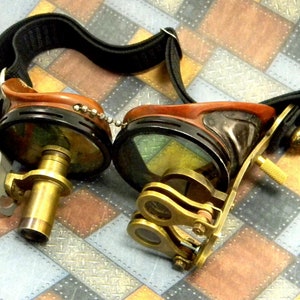 May include: A pair of steampunk goggles with brown leather accents and brass detailing. The goggles have a black strap and a chain connecting the two lenses.
