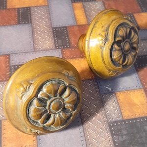 May include: Two antique brass door knobs with ornate floral designs. The knobs have a rounded shape and a raised center with a smaller circle in the middle. The knobs are on a patterned background.