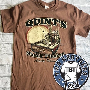 Puede incluir: Camiseta marrón con un gráfico de estilo vintage de un barco con el texto "Quint's Shark Fishing Amity Island" y un logotipo para "Two Brothers Tees".