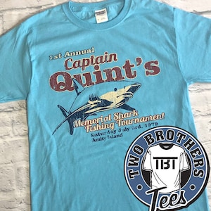 Puede incluir: Una camiseta azul claro con un gráfico desgastado de un tiburón y el texto "1st Annual Captain Quint's Memorial Shark Fishing Tournament Saturday July 3rd, 1979 Amity Island". La camiseta también tiene un logotipo azul y blanco con el texto "Two Brothers Tees" y un gráfico de una camiseta con el texto "TBT".