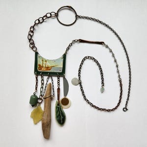 May include: A handcrafted necklace featuring a central pendant with a ship illustration, accented by various charms. The necklace includes a wooden element, green beads, and leaf-shaped details, all suspended from a chain.