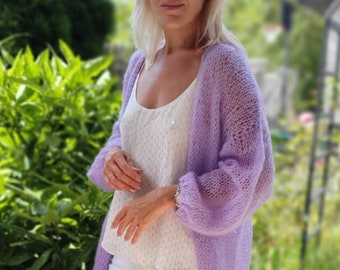 May include: A woman wearing a light purple knitted cardigan with a white top and white pants. She is standing in front of a green bush.