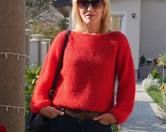 Könnte beinhalten: Eine Frau trägt einen roten Strickpullover mit einem braunen Gürtel und dunkelblaue Jeans. Sie steht vor einem Haus mit einem Ziegelsteinweg.