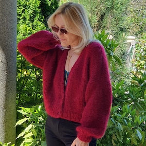 May include: A woman wearing a burgundy, oversized, hand-knitted cardigan with long sleeves. The cardigan is paired with a teal top and black pants. The background features lush green foliage and a stone pillar.