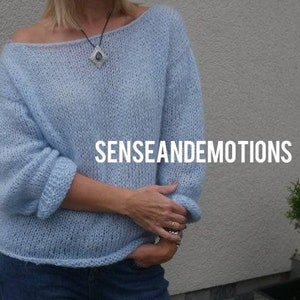 Op de afbeelding: Een lichtblauwe gebreide trui met een losse pasvorm en een licht off-shoulder halslijn. De trui is gemaakt van zacht, pluizig garen en heeft een ontspannen, casual stijl. De tekst "SENSEANDEMOTIONS" is zichtbaar op de trui.