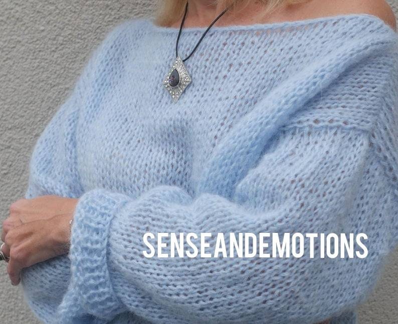 Op de afbeelding: Een lichtblauwe gebreide trui met een losse pasvorm en een brede halslijn. De trui heeft een delicate, luchtige textuur en is gemaakt van een zacht, lichtgewicht garen. De trui is versierd met een zilveren ketting met een hanger in de vorm van een traan.