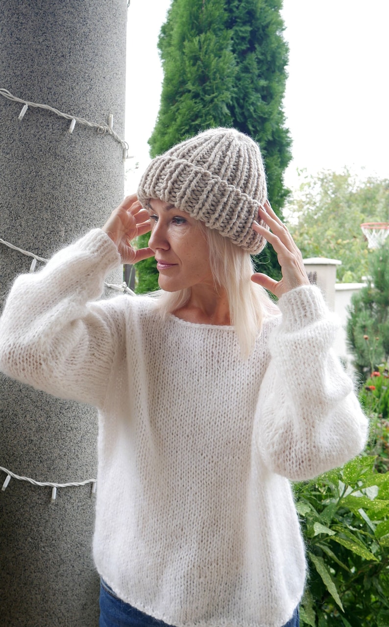 May include: A woman wearing a white knitted sweater and a beige knitted beanie hat.