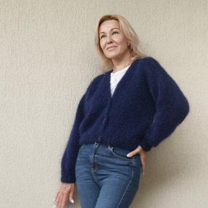 May include: A woman wearing a dark blue fuzzy cardigan with a v-neck and long sleeves. She is standing in front of a light beige wall.