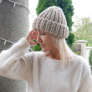 May include: A woman wearing a beige knitted beanie and a white knitted sweater.