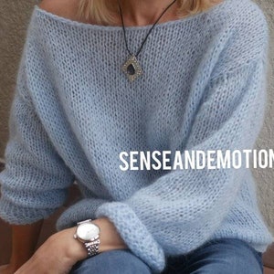 Op de afbeelding: Een lichtblauwe gebreide trui met een losse pasvorm en lange mouwen. De trui heeft een brede halslijn en is gemaakt van een zacht, pluizig garen. Het model draagt een zilveren horloge en een ketting met een donkere stenen hanger. De tekst "SENSE AND EMOTIONS" is zichtbaar op de trui.