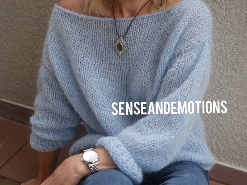 Op de afbeelding: Een lichtblauwe gebreide trui met een losse pasvorm en lange mouwen. De trui heeft een brede halslijn en is gemaakt van een zacht, pluizig garen. Het model draagt een zilveren horloge en een ketting met een donkere stenen hanger. De tekst "SENSE AND EMOTIONS" is zichtbaar op de trui.