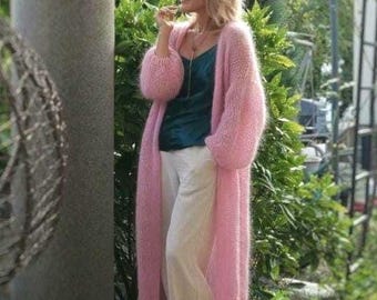 May include: A long, fuzzy, light pink open-front cardigan with long sleeves, worn over a dark teal camisole and light-colored trousers. The knit garment features a soft, textured appearance and falls below the knees, providing a layered look.