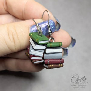 May include: A pair of silver dangle earrings with miniature stacks of books made from polymer clay. The books are in various colors, including green, red, brown, and white. The earrings are a fun and whimsical way to show your love of reading.