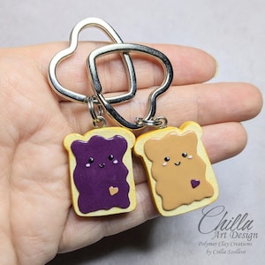 May include: Keyrings with miniature, smiling slices of toast, one with purple jam and a heart, the other with peanut butter and a heart. The keyrings are connected by a silver heart-shaped ring. The text "Chilla Art Design" is visible.