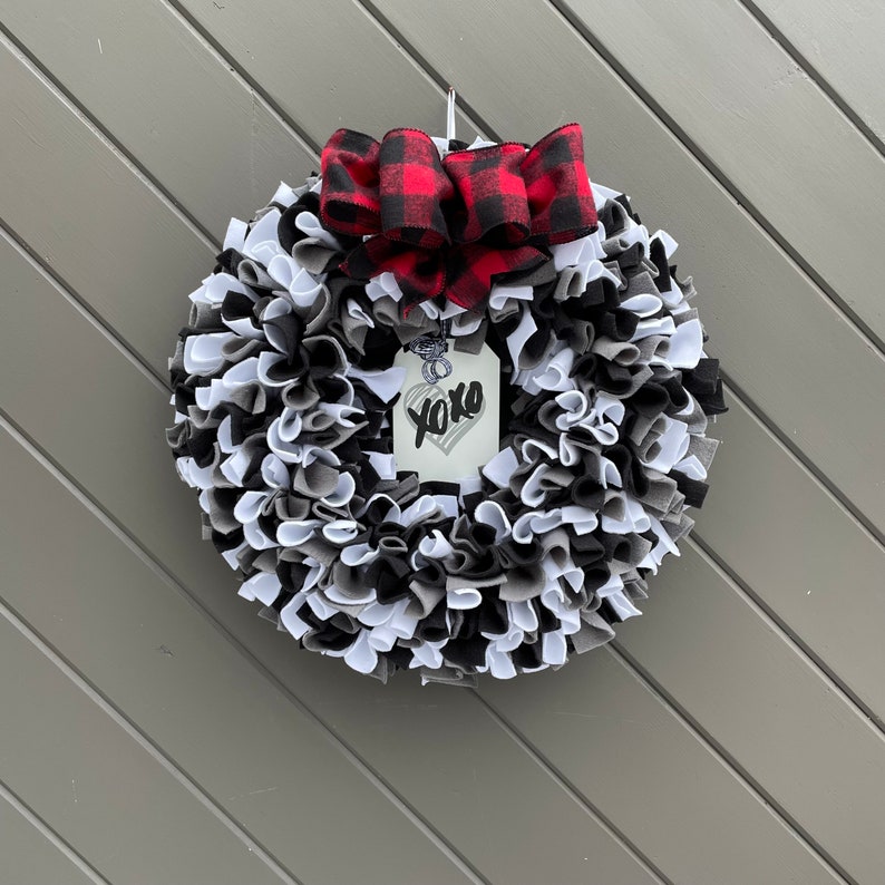 May include: A round wreath with alternating black, white, and gray fabric strips. A red and black buffalo plaid bow sits at the top. A tag with a heart and the word "xoxo" hangs from the center.