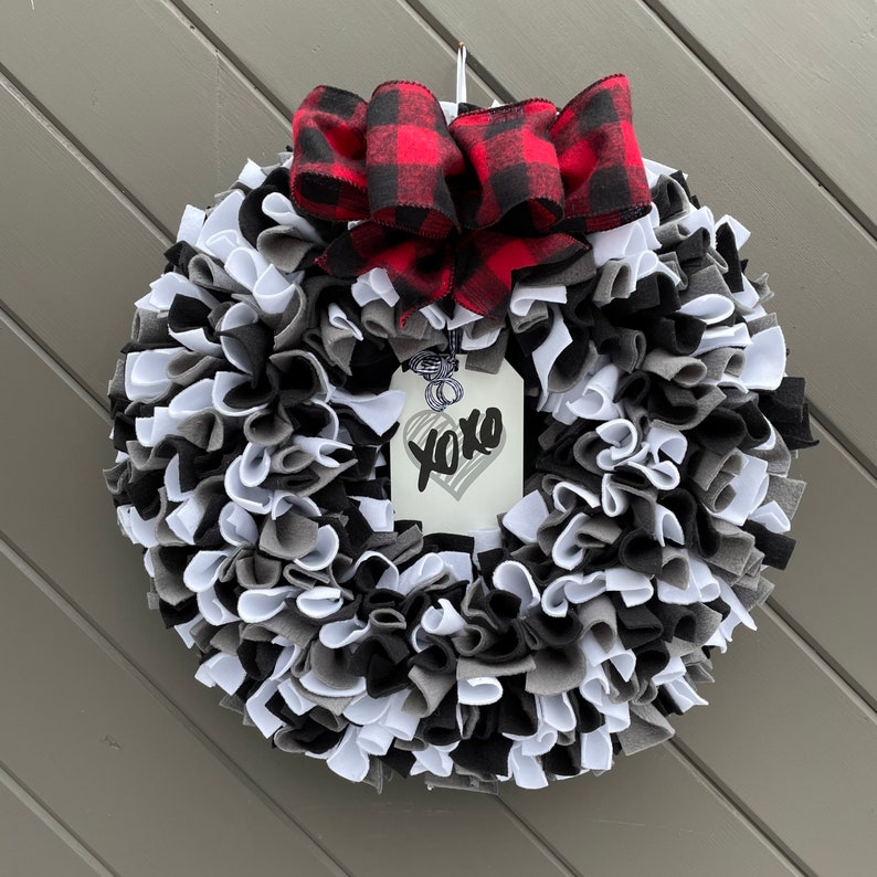 May include: A handmade wreath with a mix of black, white, and gray fabric strips. A red and black buffalo plaid bow sits at the top. A tag with a heart and the text "XOXO" hangs in the center. This decorative wreath is perfect for any door.