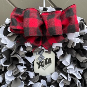May include: A handmade wreath with a red and black buffalo plaid bow. The wreath is made of white, gray, and black fabric strips. A tag with a heart and the text "xoxo" is attached to the wreath.