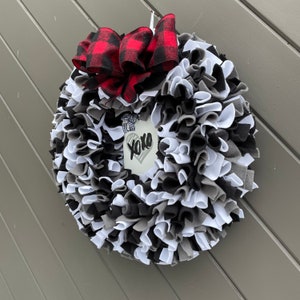 May include: A handmade wreath with a mix of black, white, and gray fabric strips. A large red and black buffalo plaid bow sits at the top. A small white tag in the center has the words "xoxo" and a heart.