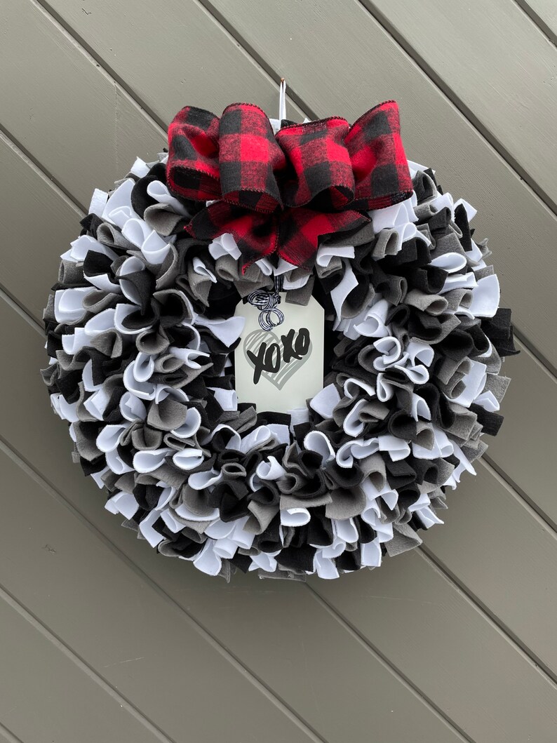 May include: A handmade wreath with a mix of black, white, and gray fabric strips. A red and black buffalo plaid bow sits at the top. A tag with "xoxo" and a heart hangs in the center.