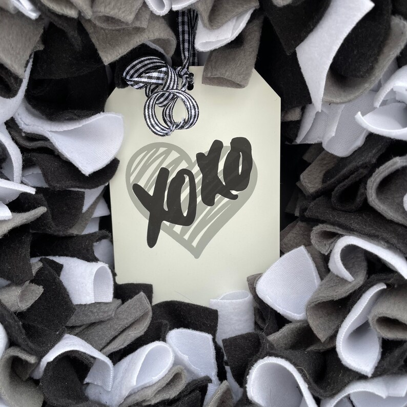 May include: A handmade wreath composed of white, gray, and black fabric strips. A cream-colored tag with the word "xoxo" in black script and a gray heart is attached with a black and white checkered ribbon.