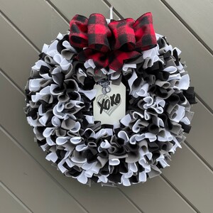 May include: A handmade wreath with a mix of black, white, and gray fabric strips. A red and black buffalo plaid bow sits at the top. A tag with "xoxo" and a heart is attached to the wreath.